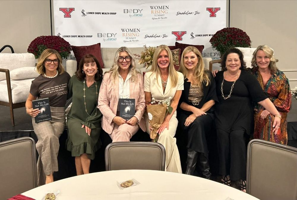 Seven women seated together on a stage in front of the Women Rising Summit banner, two of them holding copies of This Is Not My Life