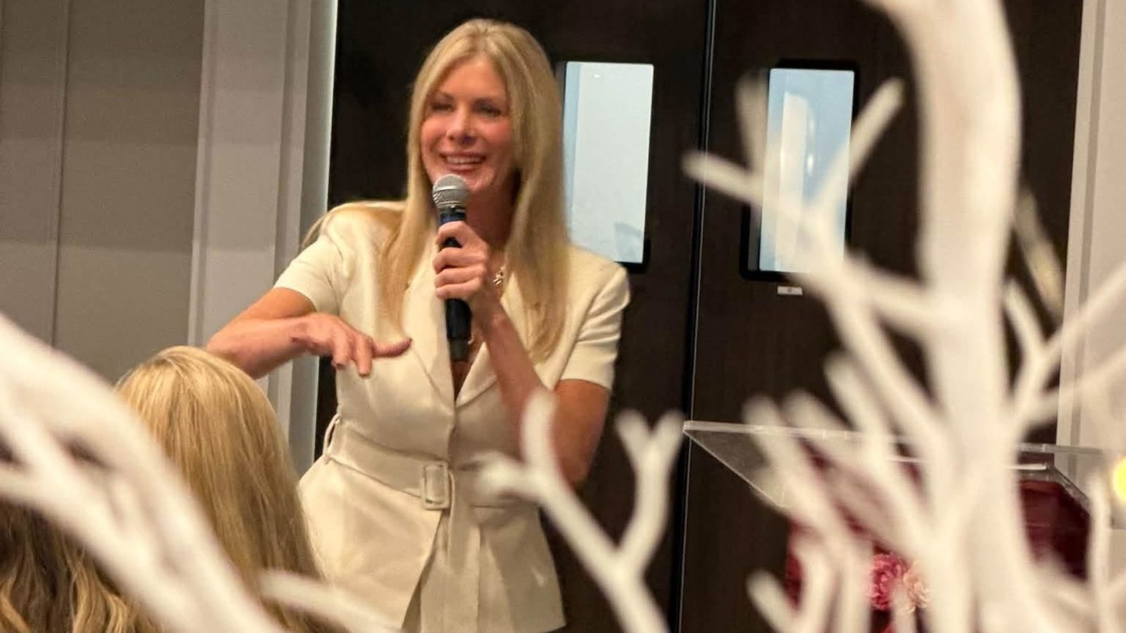 Author Laura Burkett speaking into a microphone during her keynote at the Women Rising Summit, seen through white decorative branches