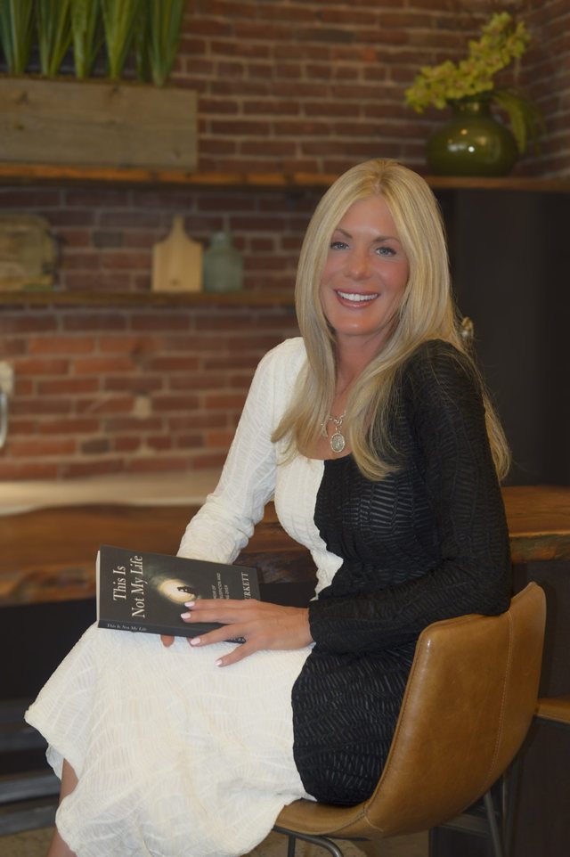 Author Laura Burkett seated in a brick-walled room, holding a copy of her memoir This Is Not My Life